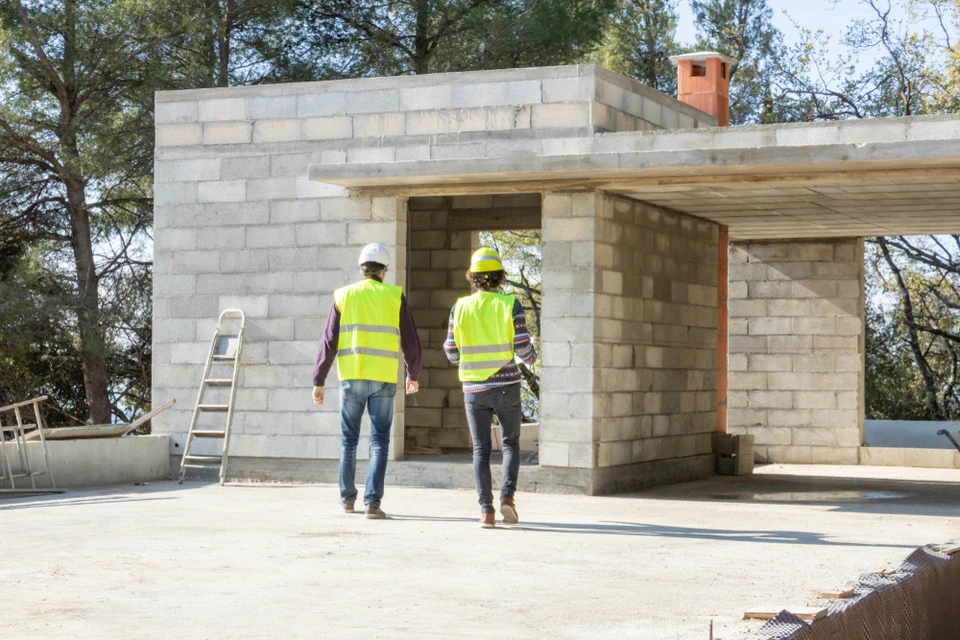 Chantier de construction extérieur avec deux ouvriers en gilets haute visibilité jaunes et casques blancs inspectant une structure en parpaings de béton