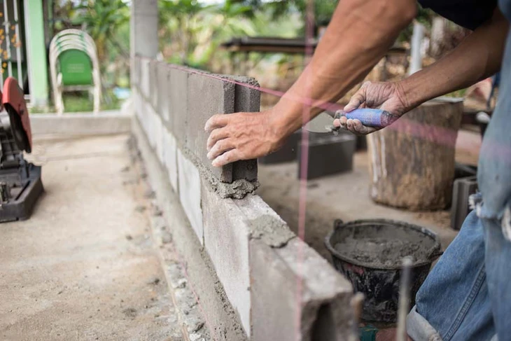 Portrait en plan rapproché ou mi-corps d'un artisan maçon d'âge mûr, cheveux gris, portant tenue de travail ou vêtements professionnels, debout sur un chantier de construction actif avec des matériaux de chantier en arrière-plan (parpaings, excavation, éléments structurels), regard vers la caméra, expression sérieuse et professionnelle reflétant 20 ans d'expérience dans le bâtiment.