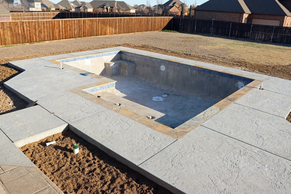 Chantier extérieur d'une piscine en béton armé en phase de construction, avec coffrage en bois et acier visible, treillis d'armature exposé, et terrain aménagé autour. La piscine est à mi-construction, clairement enterrée, illustrant une maîtrise technique sur un chantier complexe de terrassement et maçonnerie.