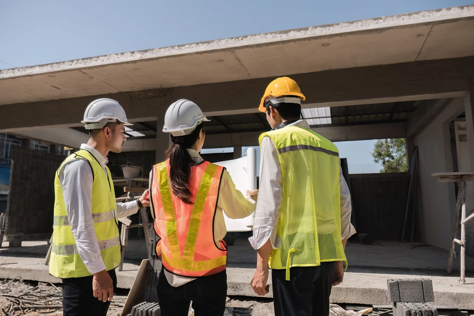 Chantier de construction résidentielle avec architecte ou contremaître en casque blanc et salopette bleue présentant des plans à un couple de clients