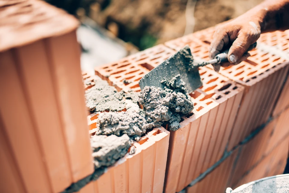 Maçon en blouson de travail examinant les plans de construction avec un client debout à ses côtés sur un chantier actif où des murs en parpaing sont visibles, montrant une interaction directe et une supervision personnelle du projet.