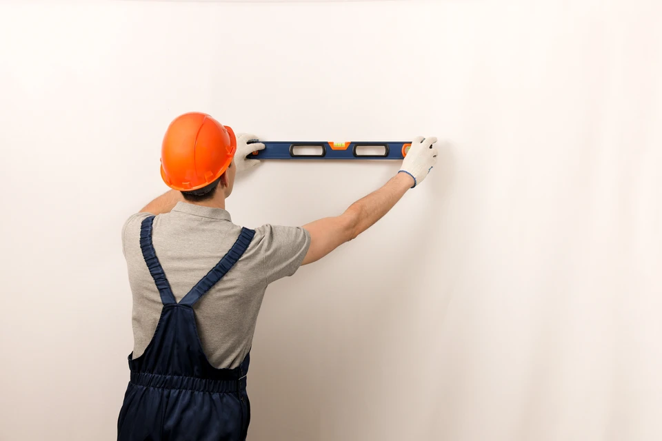 Ouvrier en salopette bleue et casque blanc utilisant un niveau à bulle jaune pour vérifier l'aplomb d'un montant de cloison sèche dans un bâtiment en construction